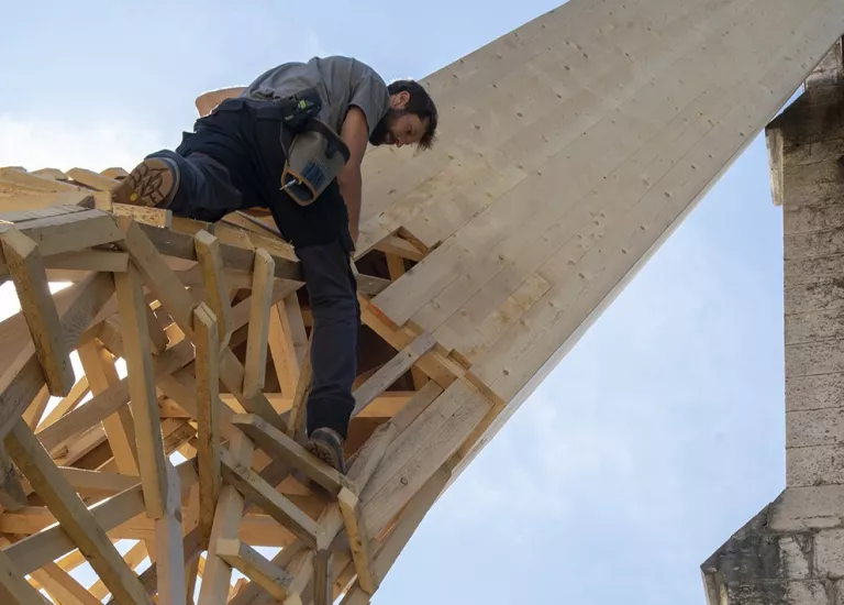 L'artiste Simon Augade assemble les chutes de bois en hauteur sur la structure de l'oeuvre "Fondement(s)"