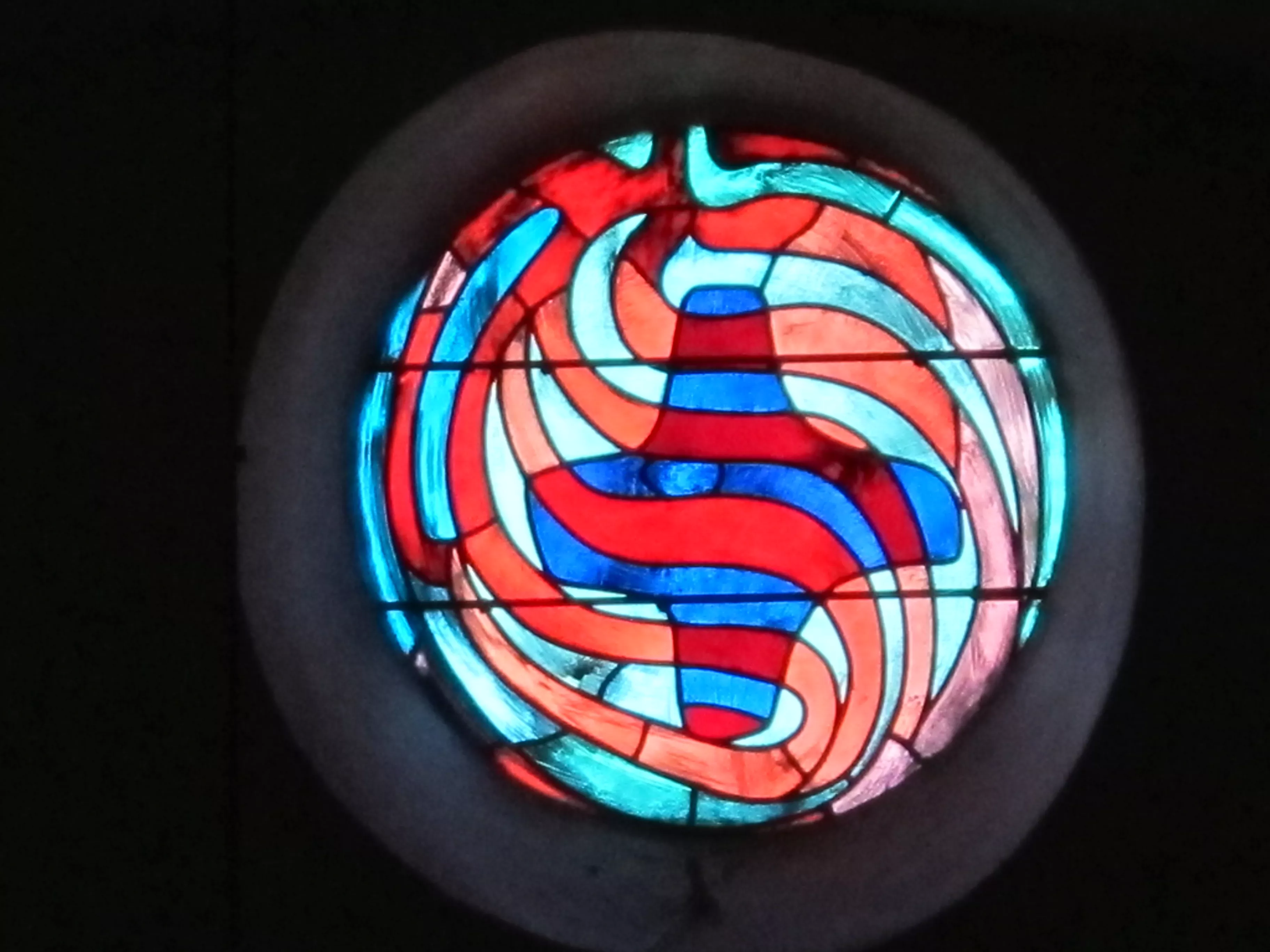 Vitrail rond avec des formes abstaites sur le thème de la vie de Jean-Baptiste. Croix abstraite bleue au centre du vitrail