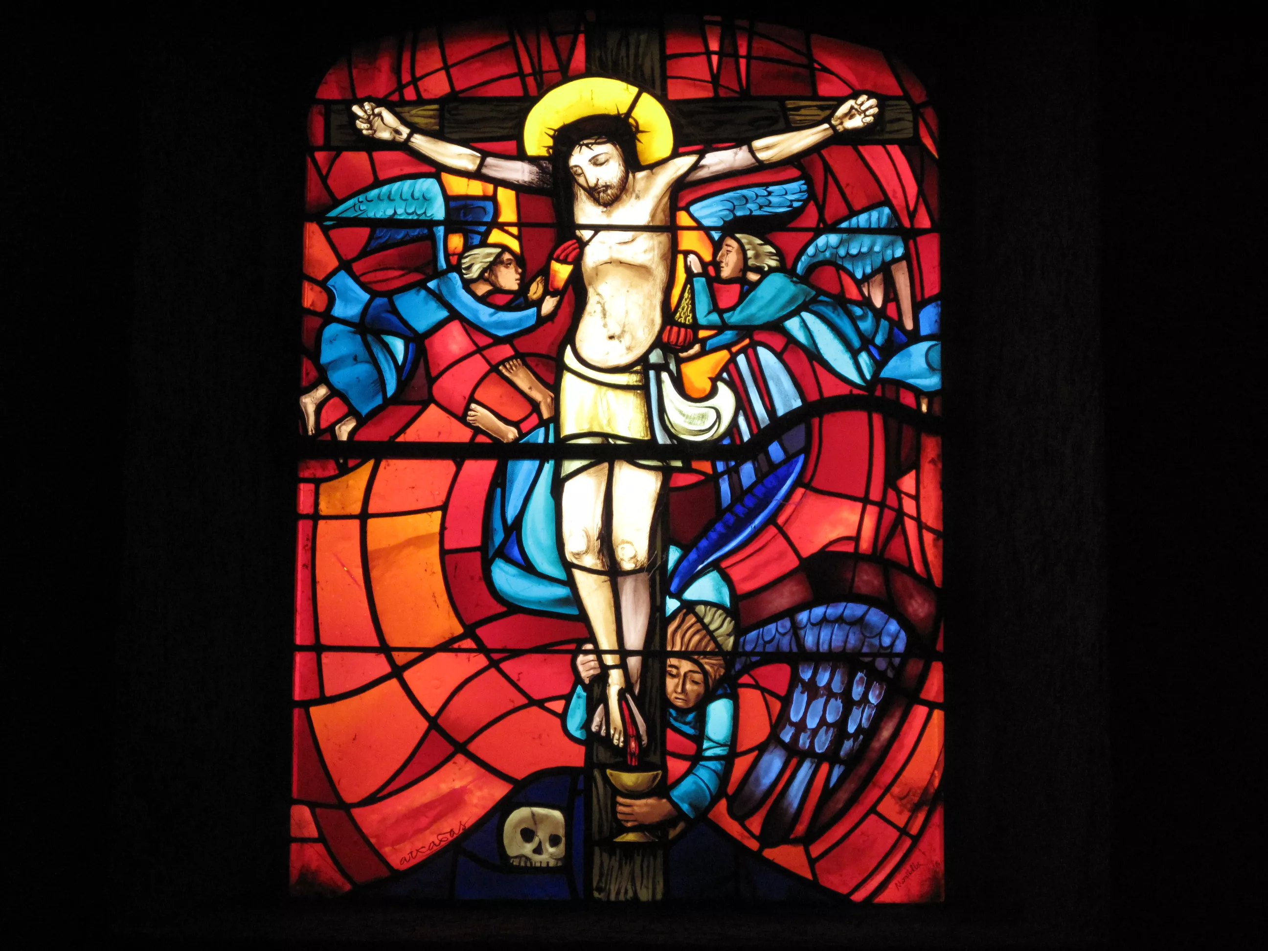 Jésus est crucifié. Deux anges bleus l'entourent au niveau du torse. Un troisième ange recueille le sang qui s'écoule des pieds du Christ. Au pied de la croix, un crâne, symbole de la vanité. Vitrail sur fond rouge.