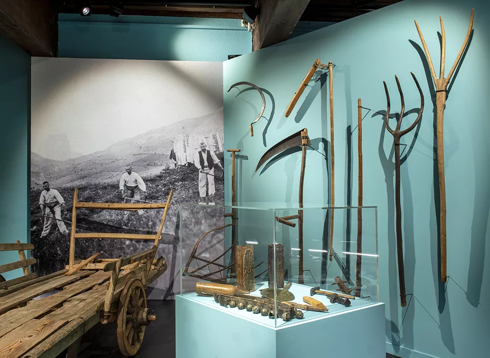 Photographie d'une salle de l'exposition &quot;Alpins&quot; présentant une charrette en bois et divers outils de travail anciens