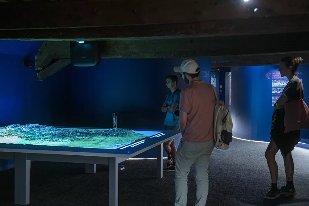 Photographie d'une salle de l'exposition &quot;Alpins&quot; présentant une carte interactive du territoire alpin observée par quelques visiteurs