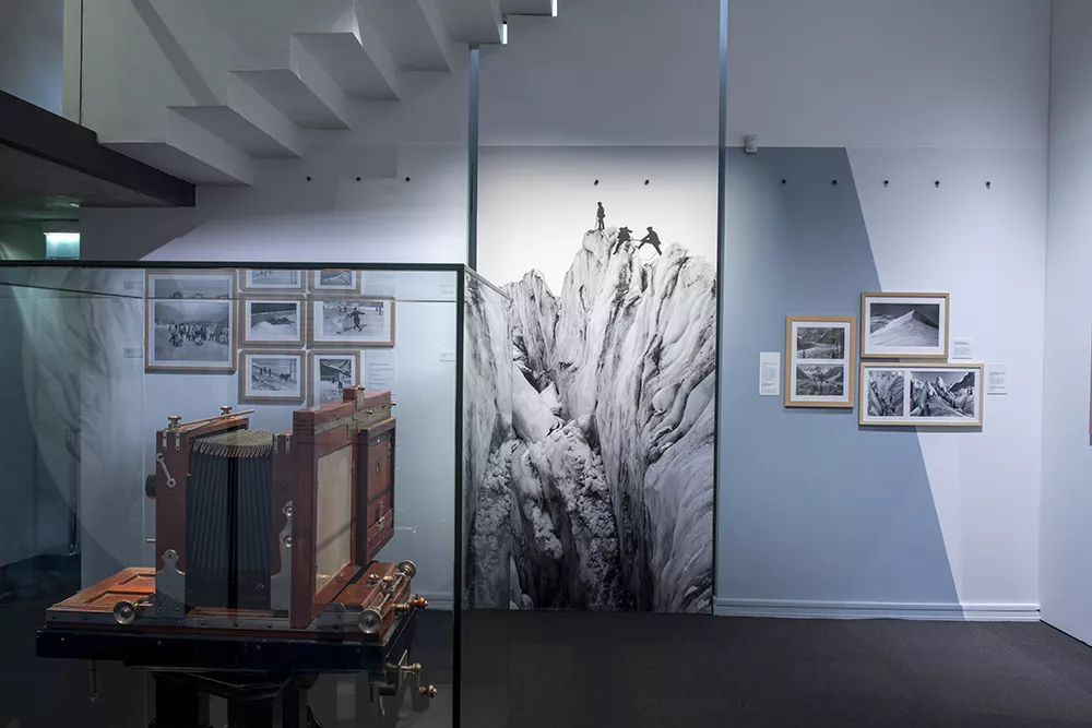 Photographie d'une salle de l'exposition &quot;Tairraz&quot; présentant des photographies de montagne en noir et blanc et une vitrine avec un appareil photo ancien