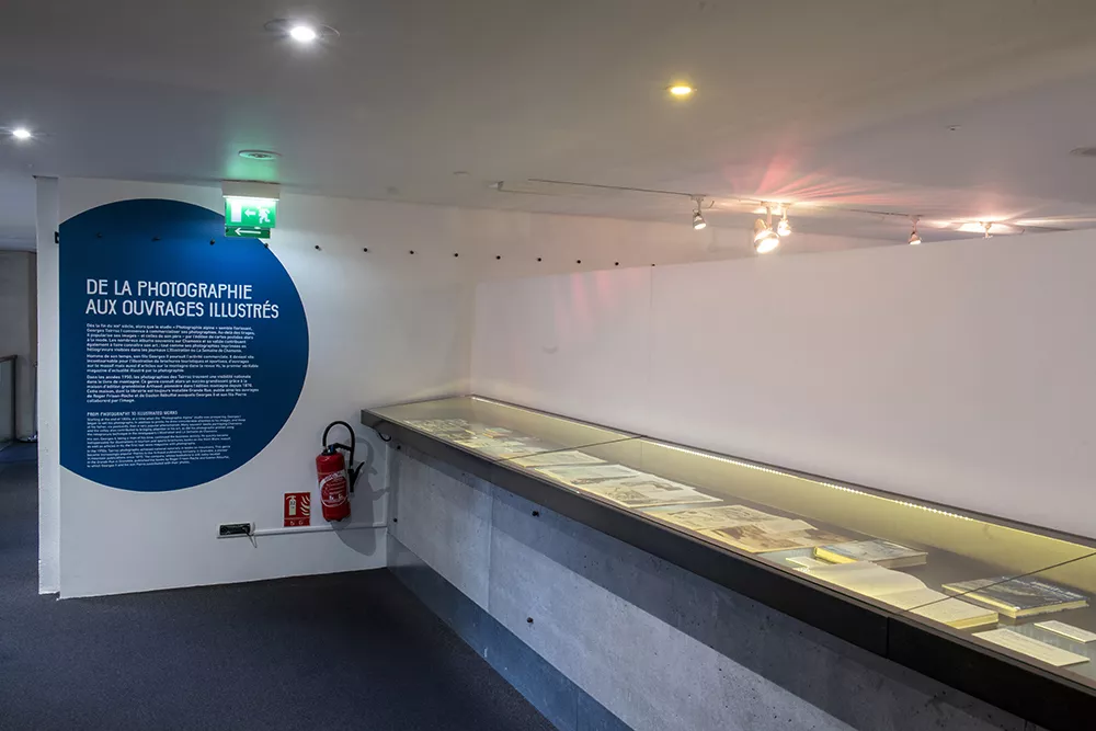 Photographie d'une salle de l'exposition &quot;Tairraz&quot; présentant une vitrine de livres et cartes postales anciennes