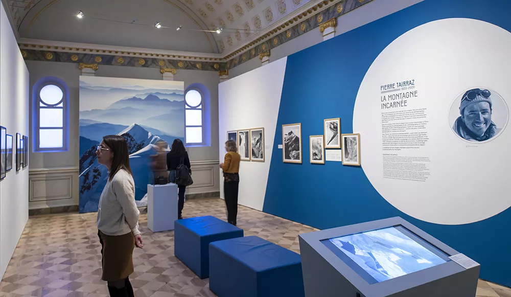 Photographie d'une salle de l'exposition &quot;Tairraz&quot; présentant des photographies de montagne en noir et blanc regardées par quelques visiteurs