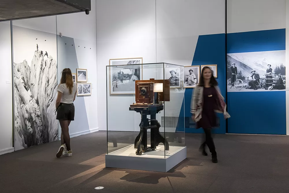 Photographie d'une salle de l'exposition &quot;Tairraz&quot; présentant des photographies de montagne en noir et blanc et une vitrine avec un appareil photo ancien