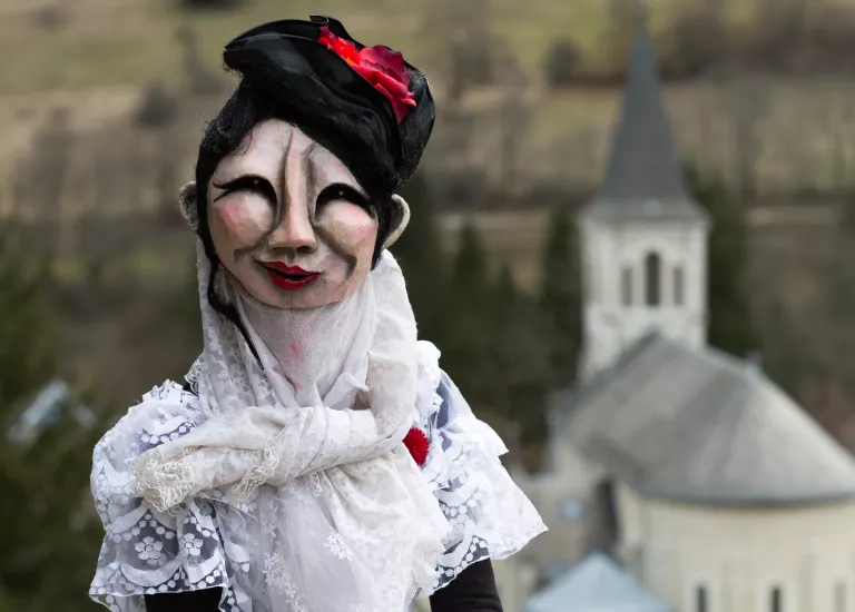 Photographie d'un personnage du Turak Théâtre masqué portant une robe de mariée devant l'église Saint Hugues © Raphael Licandro