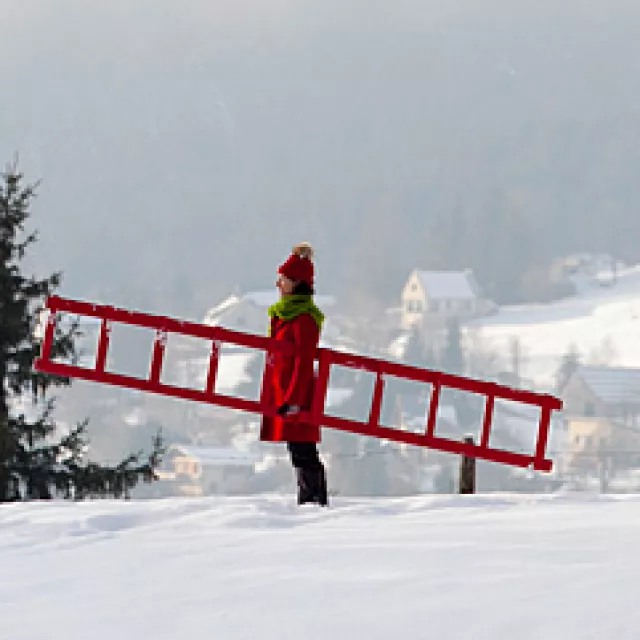 un homme et une femme habillés en rouge ou en bleu portant une échelle rouge dans un paysage de neige © Stéphanie Nelson