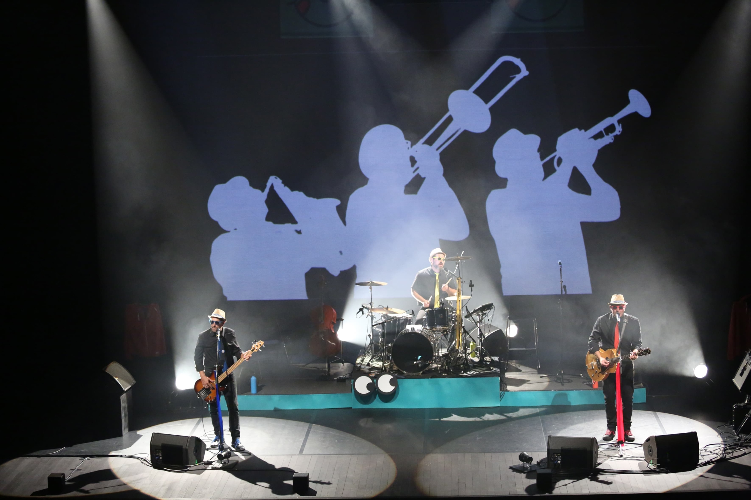 Concert des Méli Mômes au MRDI ! © Yannick Perrin Photographie d'une scène de concert avec un bassiste, un batteur et un guitariste, et une projection en fond de scène de l'ombre de trois musiciens jouant du saxophone, du trombone et de la trompette © Yannick Perrin