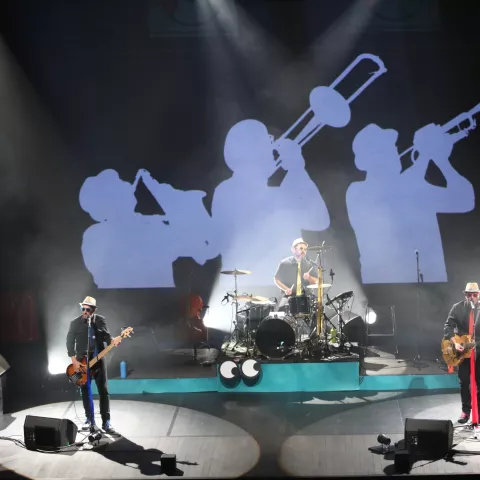 Photographie d'une scène de concert avec un bassiste, un batteur et un guitariste, et une projection en fond de scène de l'ombre de trois musiciens jouant du saxophone, du trombone et de la trompette © Yannick Perrin