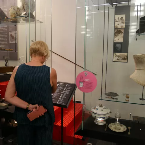 Photographie d'une salle de musée avec une femme de dos lisant les cartels explicatifs des objets présentés en vitrine