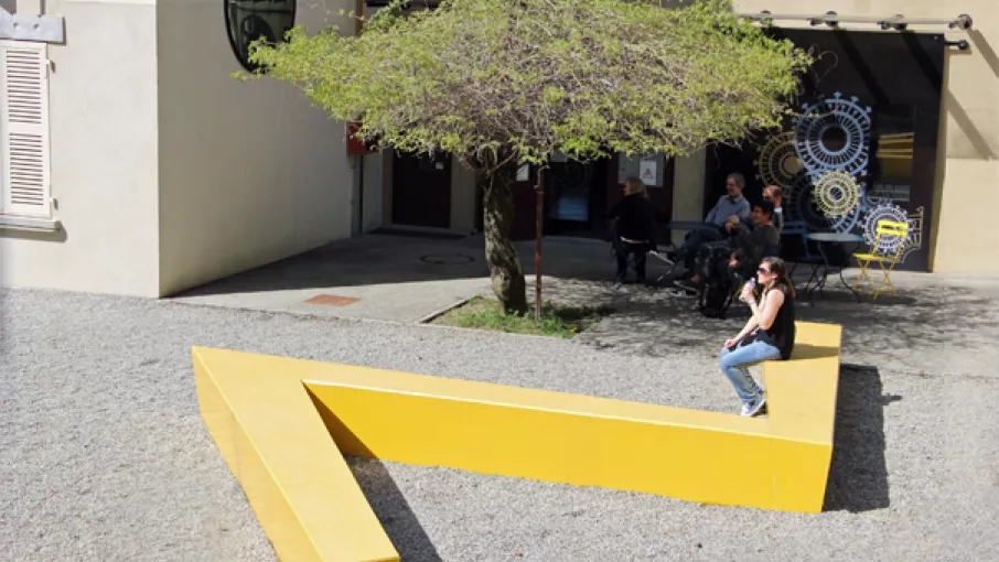 Maison Bergès à Lancey, entrée devant laquelle la glycine fait de l'ombre aux personnes assises autour des tables de jardin. Une femme est assise sur le banc en béton de couleur jaune et en forme d'éclair. © Maison Bergès, photo Frédérique Virieux